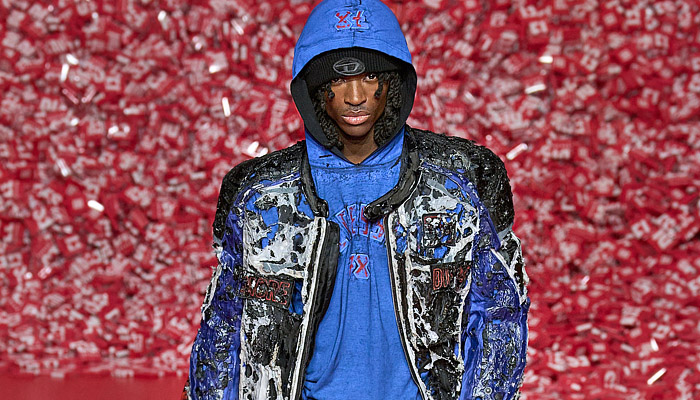 Diesel 2023-2024 Fall Autumn Winter Mens Runway Presentation - Milan Fashion Week Italy - Hyperreal Close-Up Prints Faces Smiles Teeth Pop Art Membrane Sheer Tulle Blow Torch Destroyed Destructed Peel Off Motorcycle Biker Jacket Leather Shredded Devoré Frayed Raw Hem Slouchy Baggy Loose Denim Jeans Burnt Cracked Quilted Puffer Hood Oversized Coat Blazer Mesh Holes Knit Grunge Marbled Shorts Crumpled 1DR Pod Wristlet Pouch Handbag XL Diesel Logo D-Charm Trainers Sneakers 